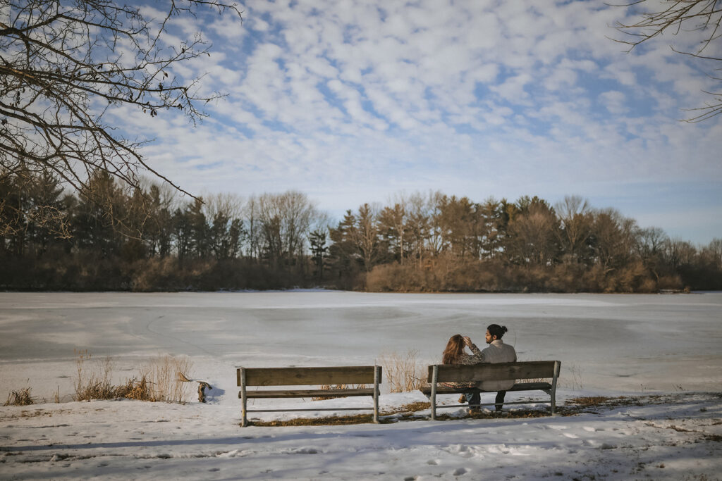 Couple sitting on a bench beside a frozen lake during a winter engagement session at Homer Lake Forest Preserve in Homer, Illinois.