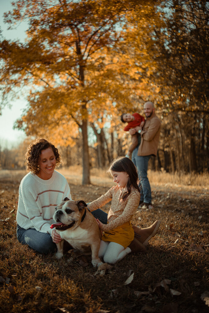 Parents with two young children and a bulldog sitting together in a scenic, fall setting at River Bend in Mahomet, IL, photographed by Light By Zoey Photography, Champaign, IL.