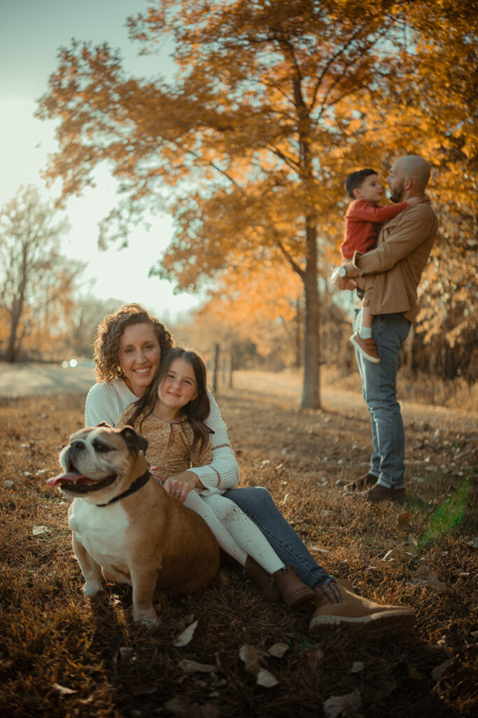 Parents with two young children and a bulldog sitting together in a scenic, fall setting at River Bend in Mahomet, IL, photographed by Light By Zoey Photography, Champaign, IL.